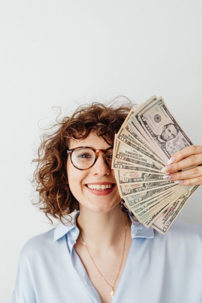 pexels-photo-7680633 Cheerful woman with curly hair and glasses holding a fan of US dollar bills.